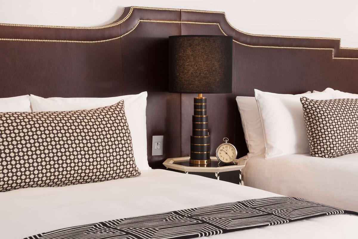 A hotel room with a large bed, two pillows with a geometric pattern, a bedside table with a lamp, and a clock.
