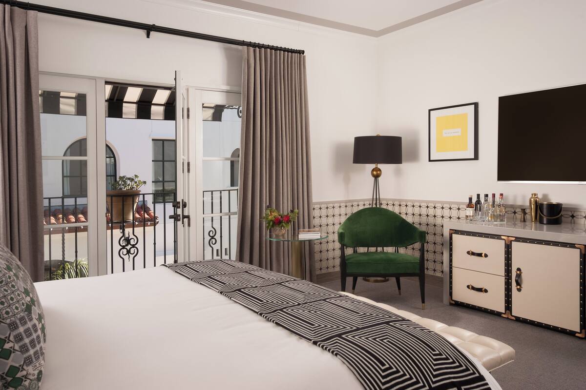 A hotel room with a bed, a green chair, a desk with drawers, a lamp, and a view of the outside.