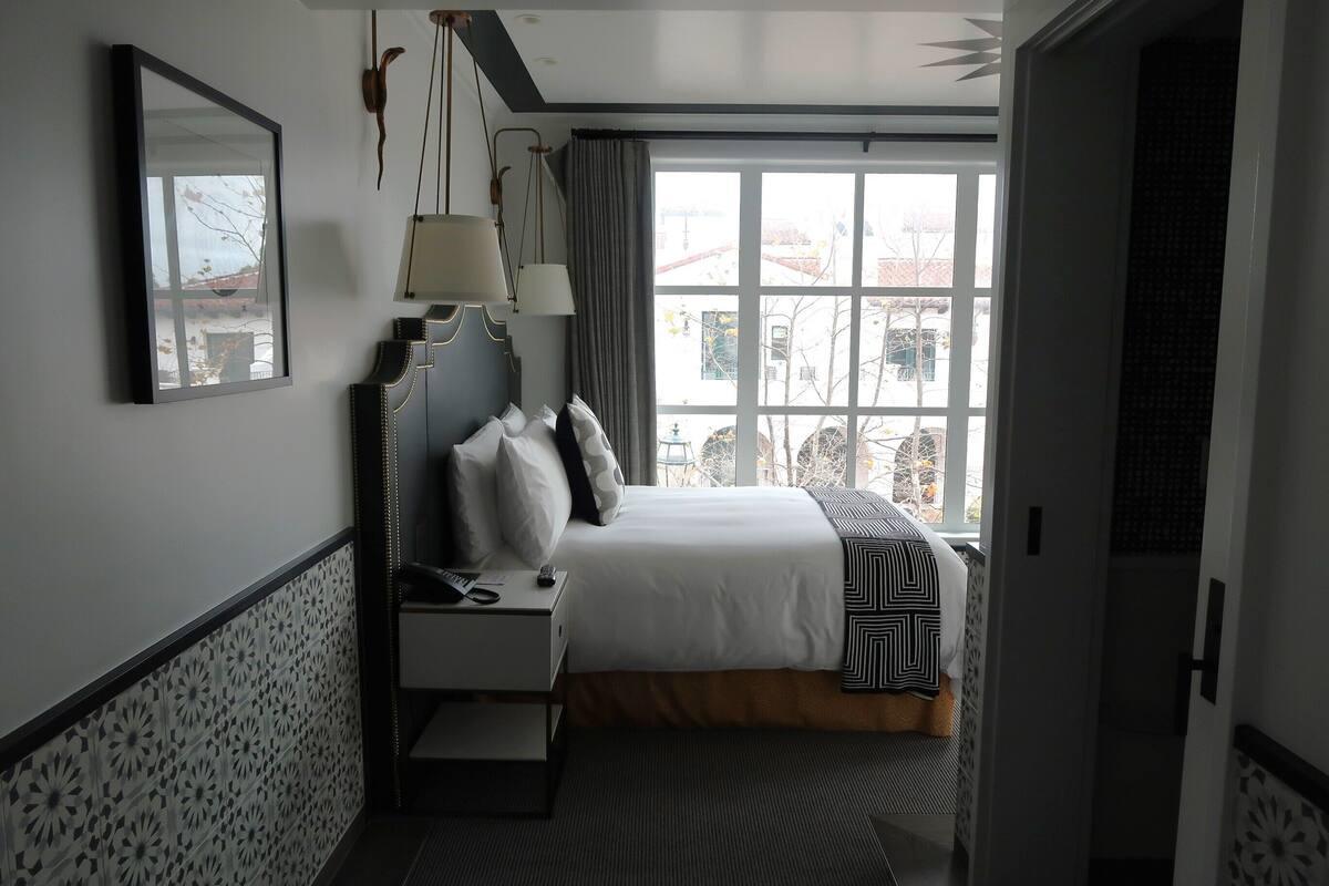 A hotel room with a bed, bedside table, lamp, and a window with a view of buildings.