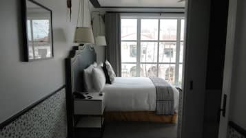 A hotel room with a bed, bedside table, lamp, and a window with a view of buildings.