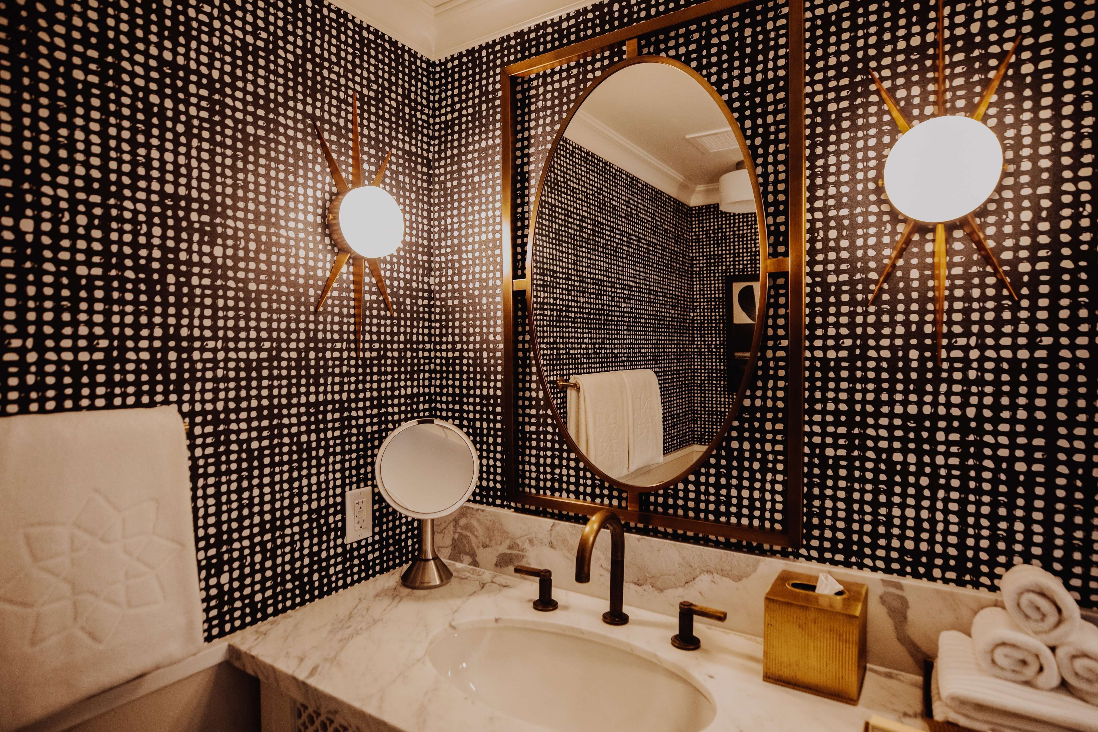 A bathroom with a marble sink, two mirrors, a towel rack, and a sun-shaped light fixture.