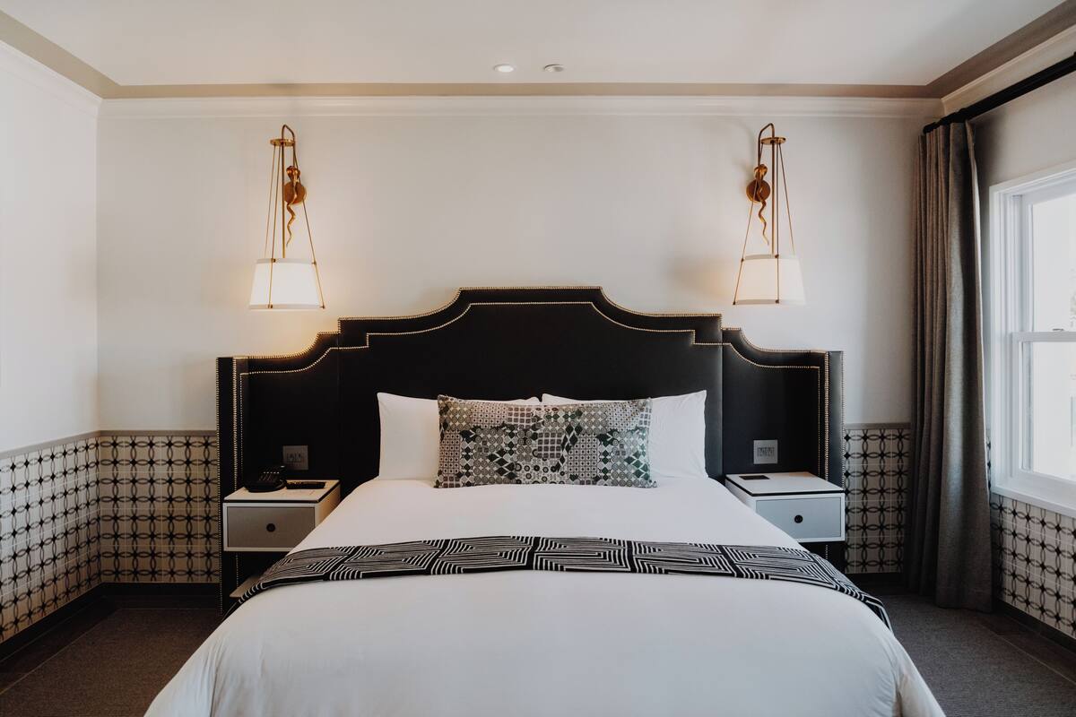 A hotel room with a large bed, two bedside tables, a patterned throw, and a window with curtains.