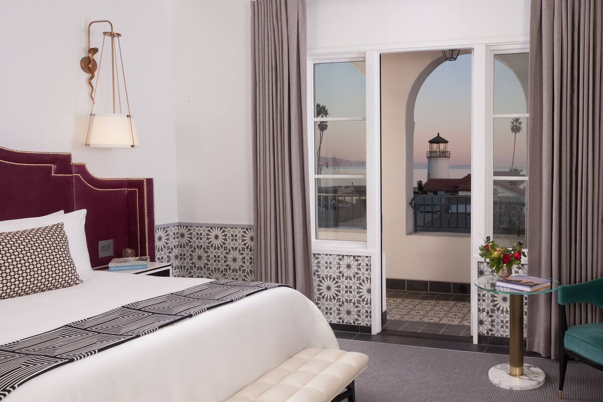 A hotel room with a bed, a bedside table, a small table with a vase of flowers, and a view of a building through a window.