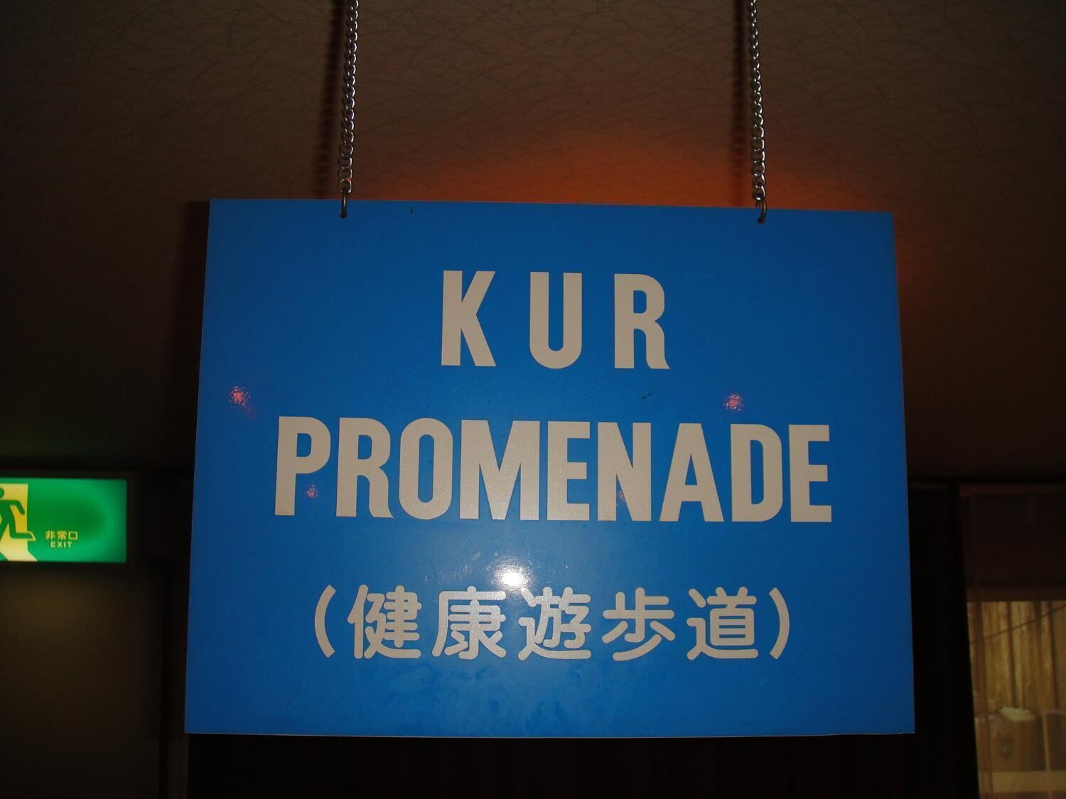 Une pancarte bleue avec du texte blanc indiquant « KUR PROMENADE (健康遶步道) » est suspendue au plafond.