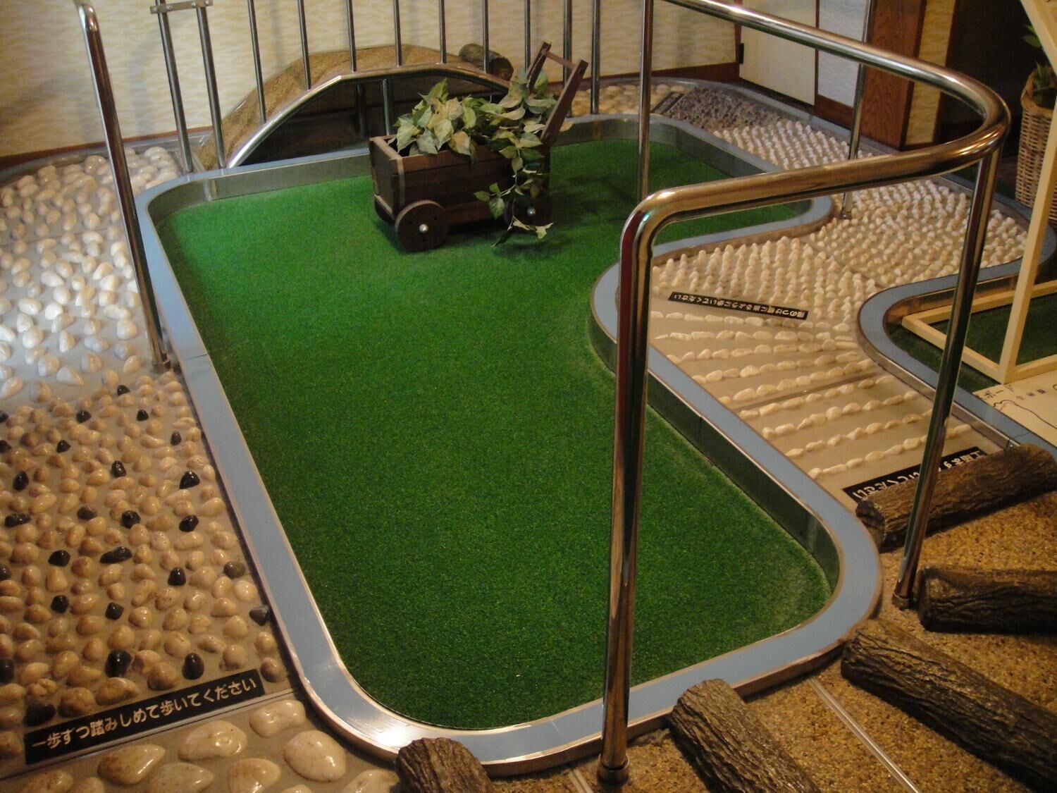 A miniature golf course with a green putting surface, wooden obstacles, and a small planter with green foliage.