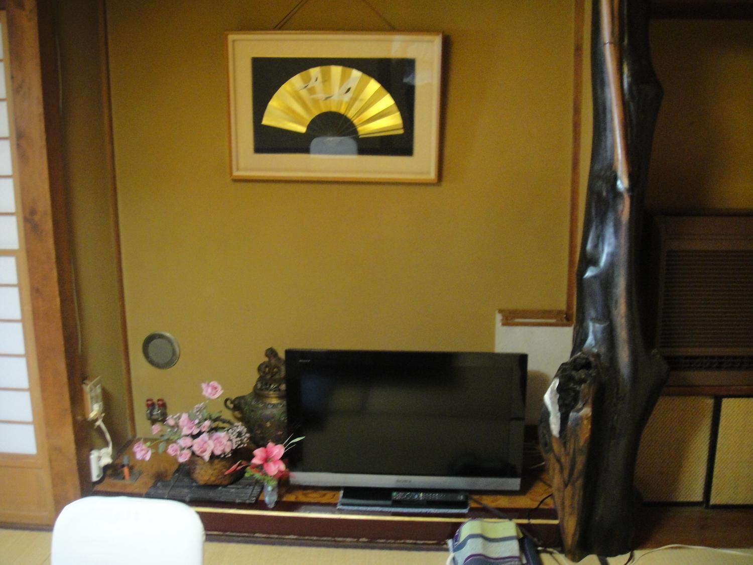 Une pièce avec un téléviseur posé sur un meuble en bois, un tableau encadré représentant un ventilateur et une petite table ornée de fleurs et d’objets décoratifs.