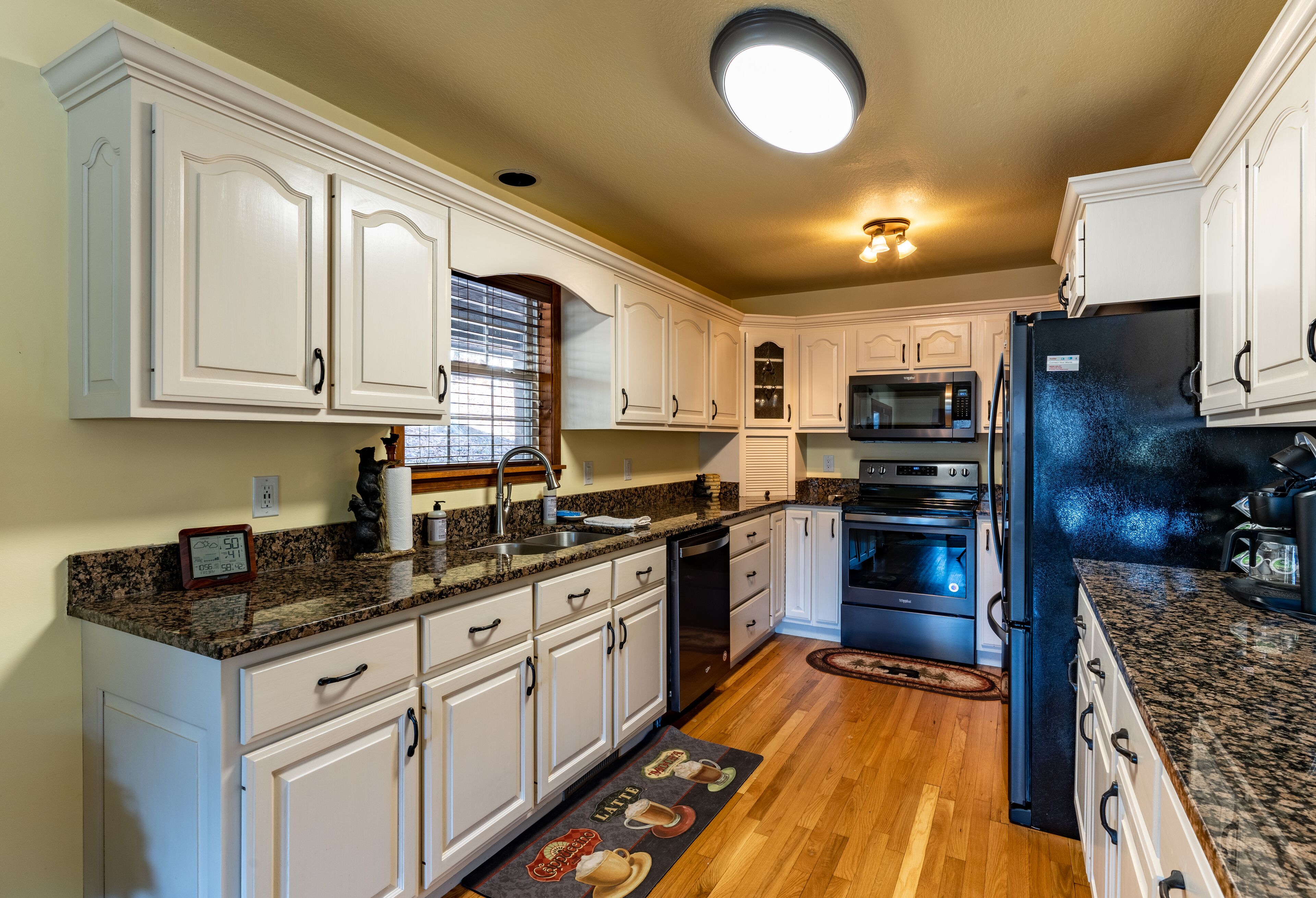 Una cocina con armarios blancos, encimeras de granito, microondas integrado, refrigerador y piso de madera.