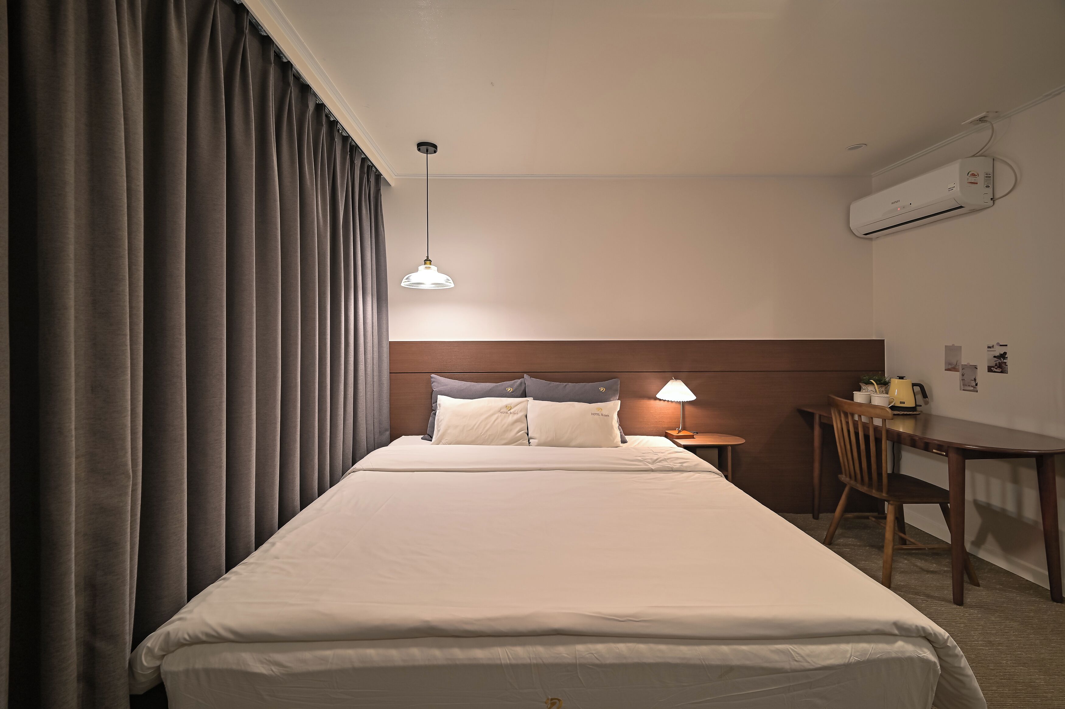 A hotel room with a large bed, a desk, a chair, a lamp, and an air conditioning unit.