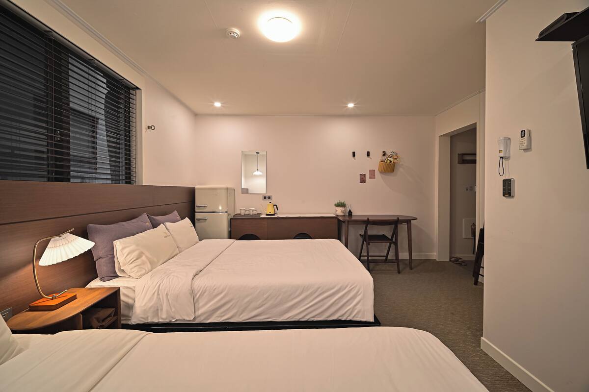 A hotel room with two beds, a desk, a chair, a lamp, a mirror, and a small table with a plant.