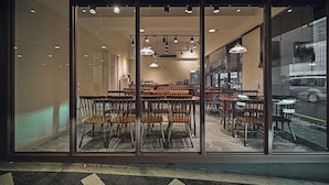 Un restaurante con una decoración moderna, que cuenta con mesas y sillas de madera, iluminación colgante y amplios ventanales con vistas a la calle.