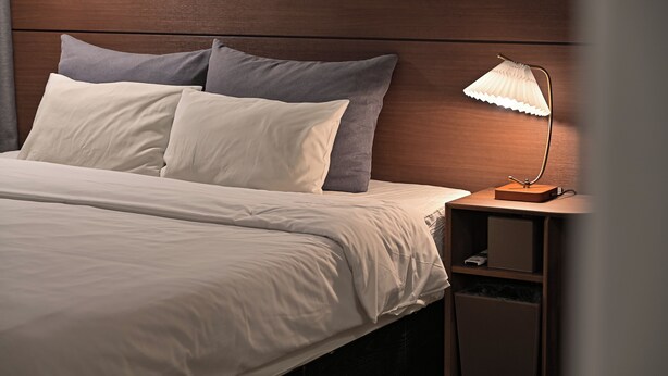 Una cama bien hecha con ropa de cama blanca y gris, un cabecero de madera y una mesita de noche con una lámpara.