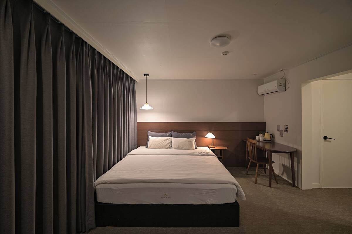 A hotel room with a large bed, a bedside table with a lamp, a small desk, and a door.