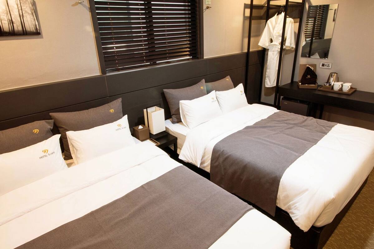 A hotel room with two beds, a nightstand, a mirror, a window with blinds, and a small table with a coffee maker and cups.