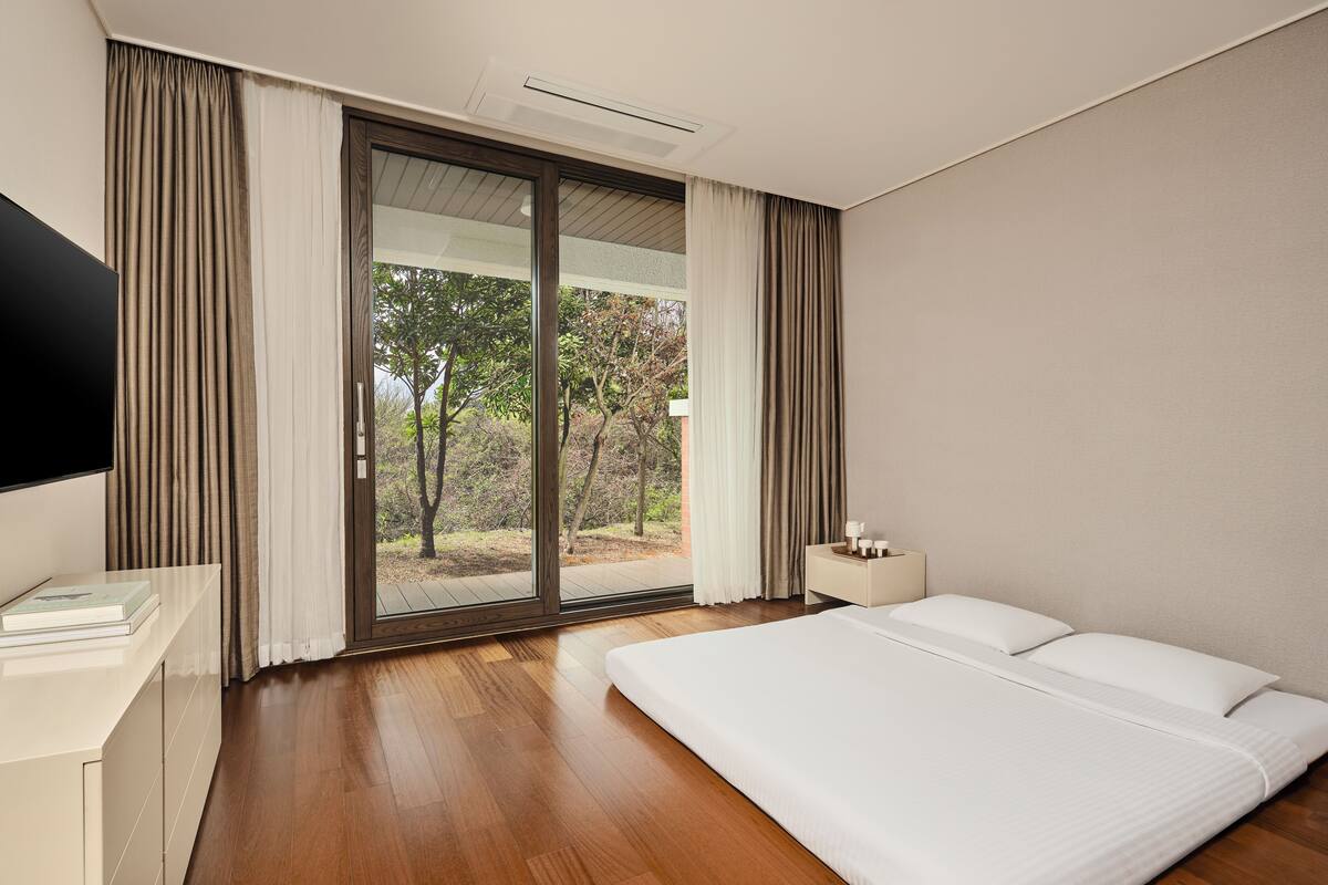 Een moderne hotelkamer met een groot bed, een flatscreen televisie, een nachtkastje en uitzicht op het omliggende landschap door een glazen schuifdeur.