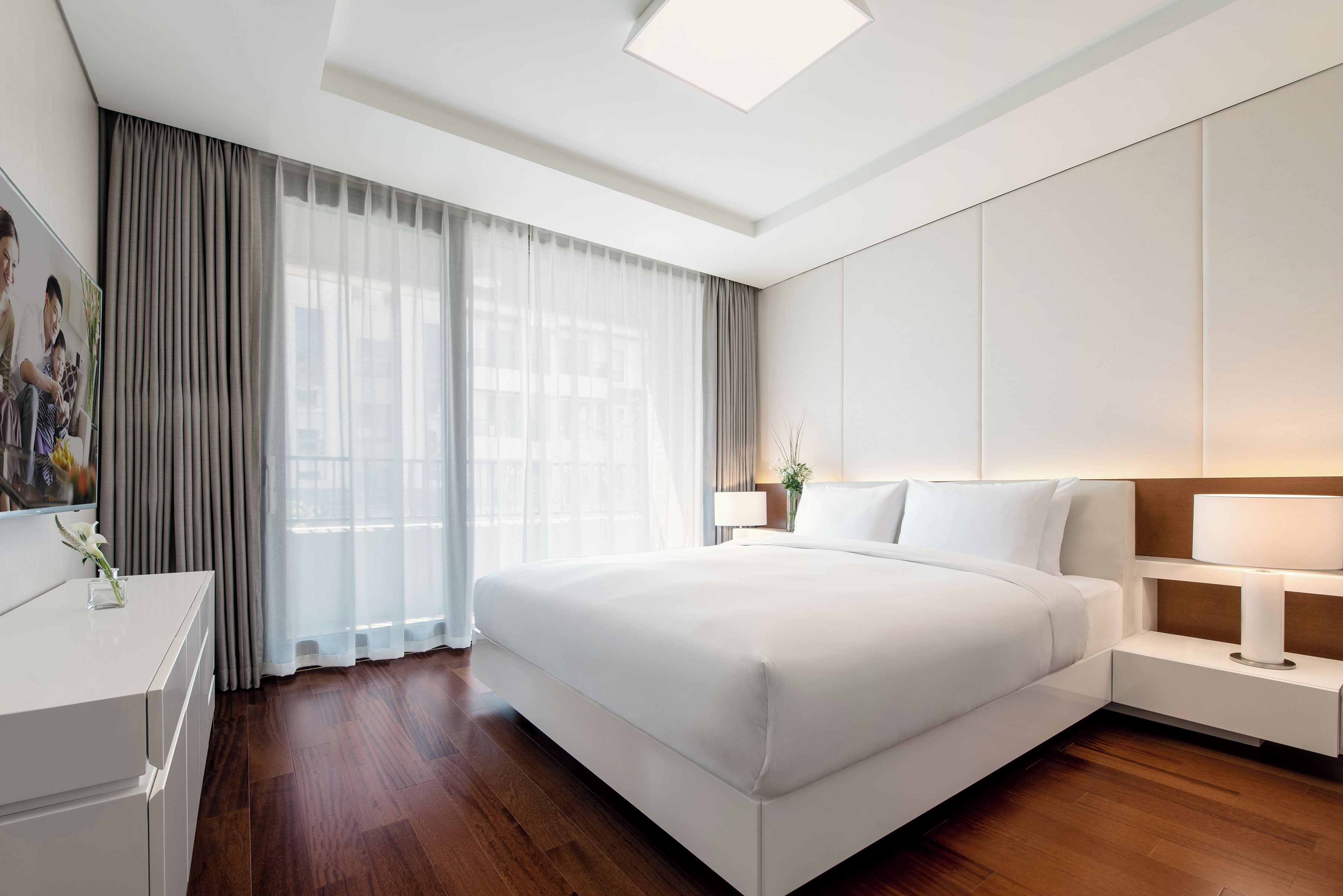 A modern hotel room with a large bed, a bedside table, a nightstand, and a flat-screen TV mounted on the wall.