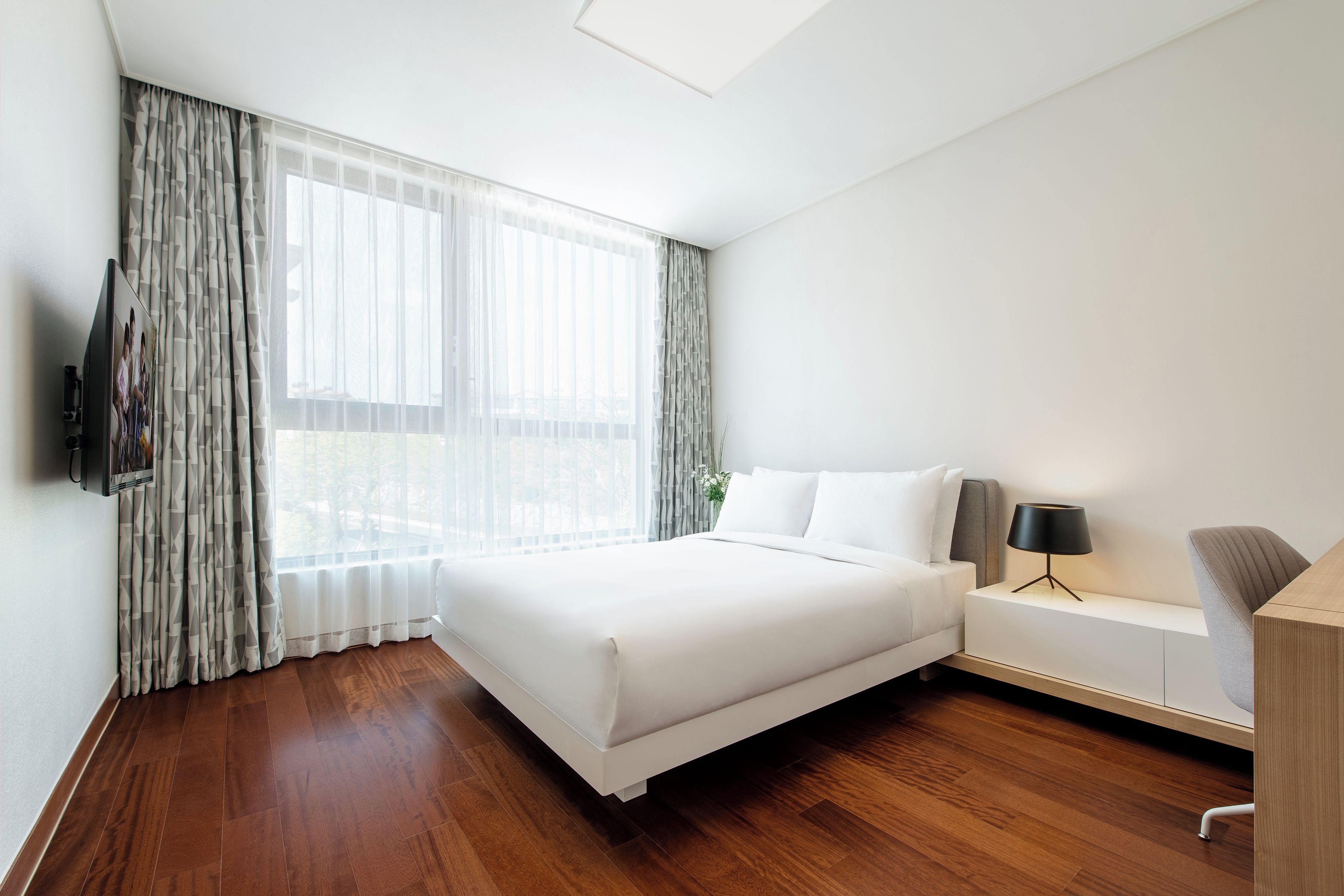 A modern hotel room with a large bed, a bedside table with a lamp, a TV mounted on the wall, and a window with sheer curtains.