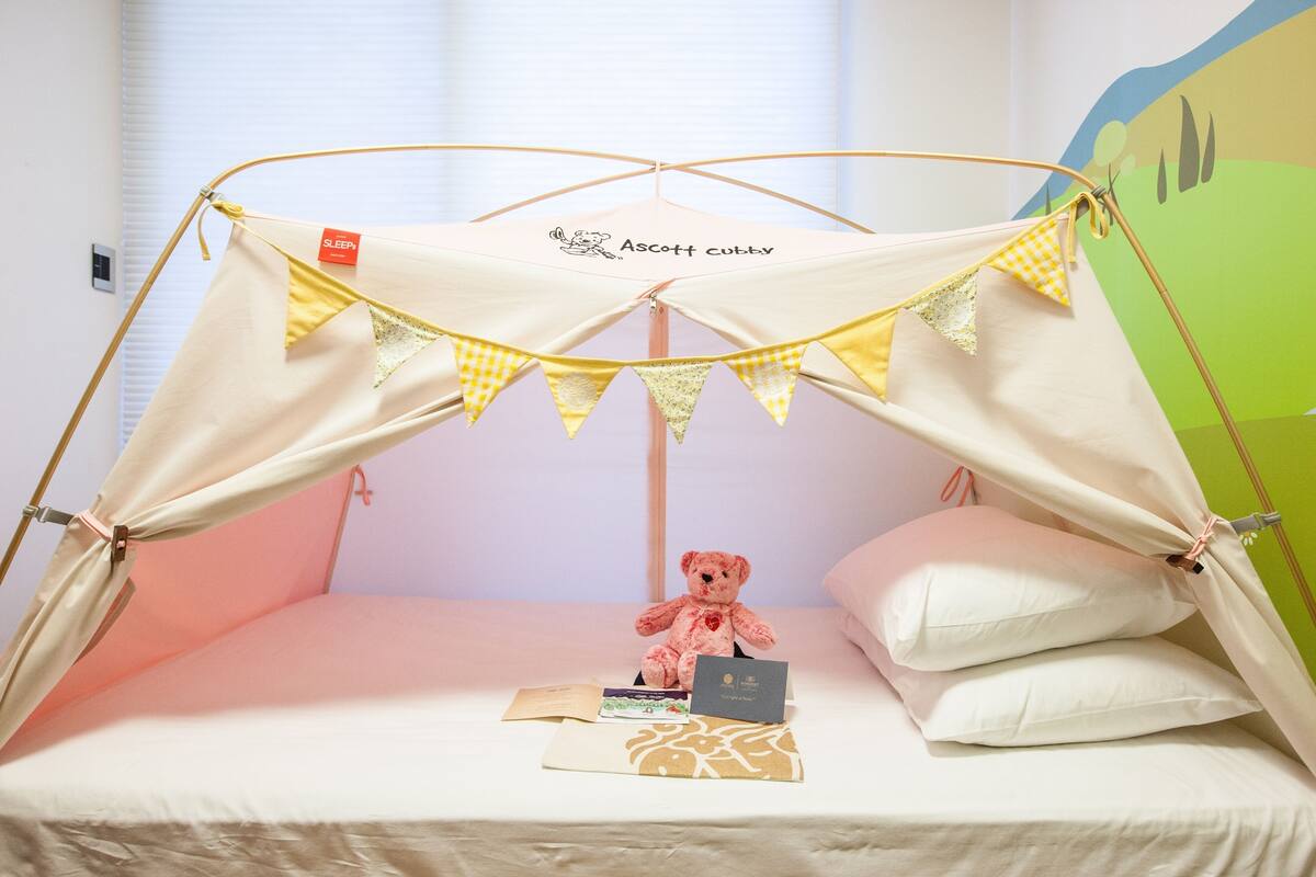 Een eenpersoonsbed met hemel, een teddybeer, en een klein tafeltje met een boek en een kaart.