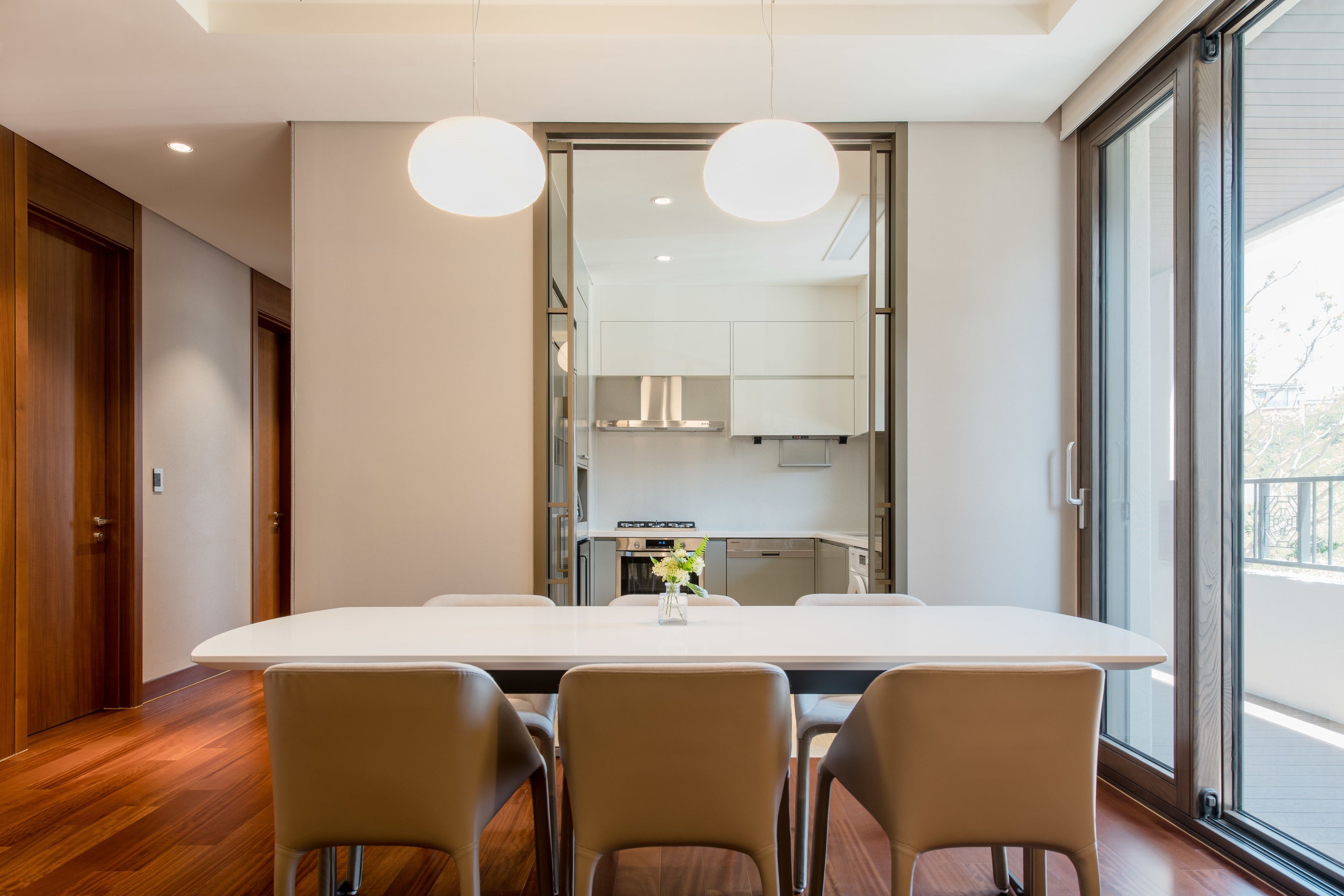 A modern dining area with a large table and four chairs, a kitchen in the background, and large windows.