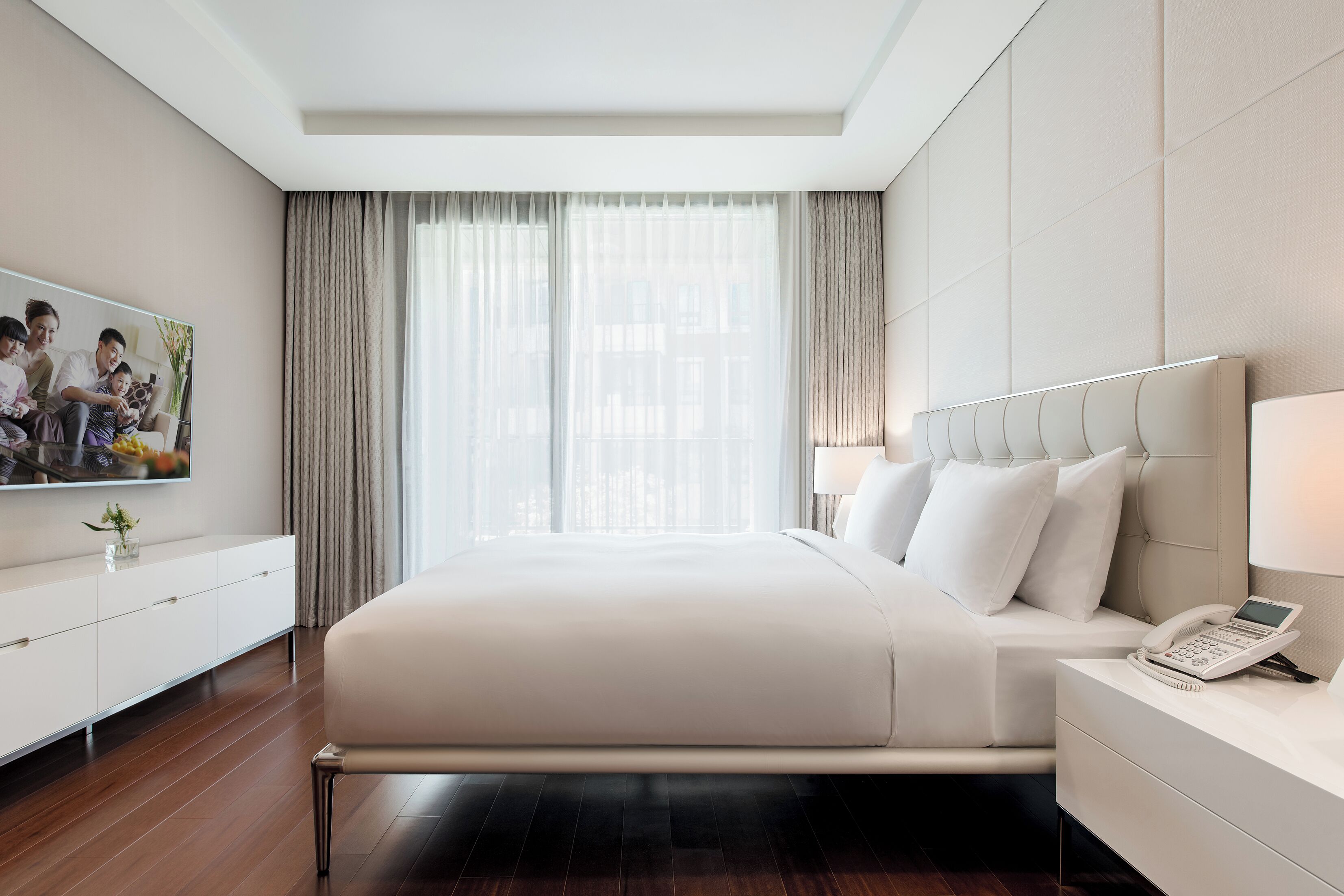 A modern hotel room with a large bed, a bedside table with a phone, a TV mounted on the wall, and a window with curtains.