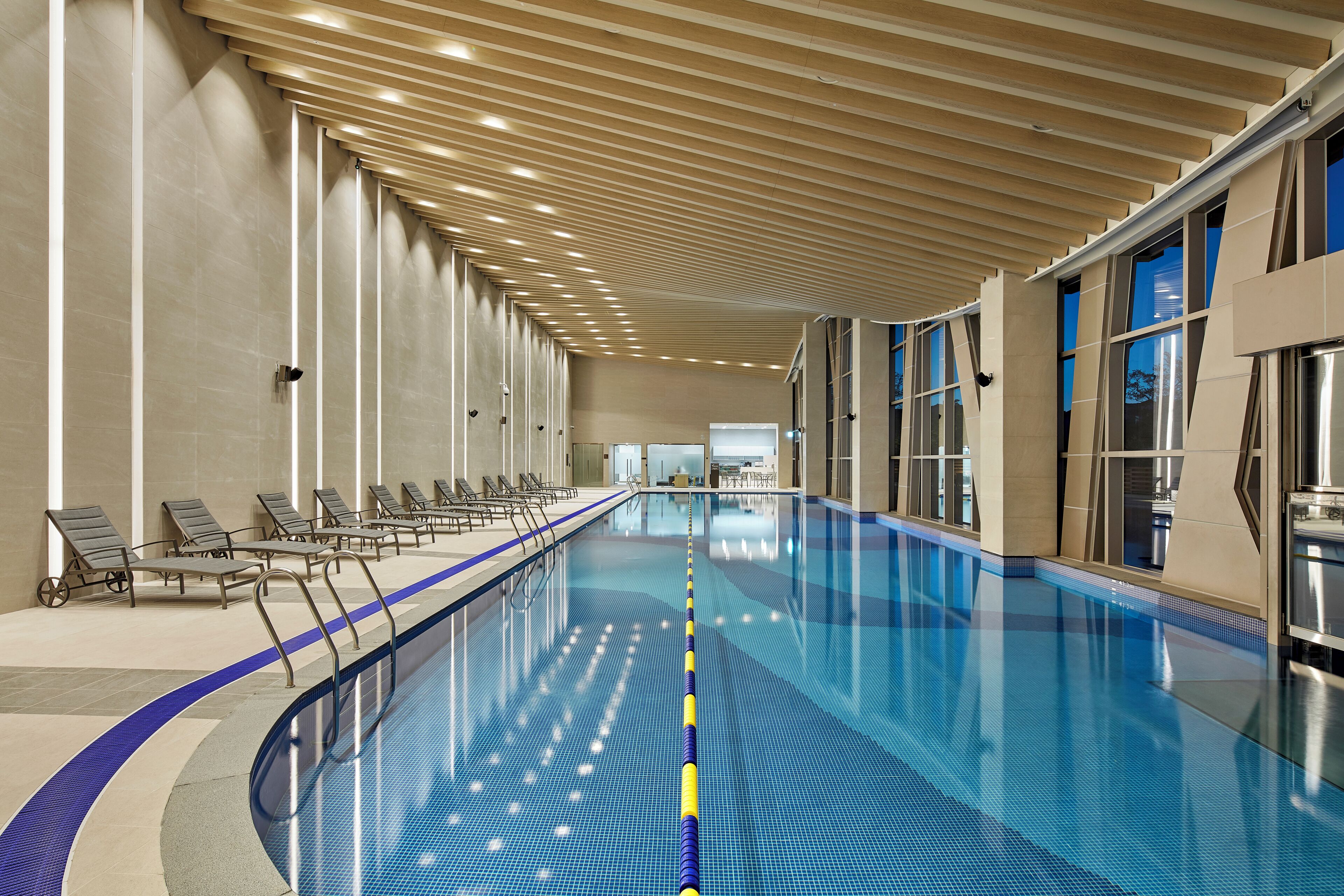 An indoor swimming pool with a long, straight layout, a tiled floor, and a ceiling with evenly spaced lights.