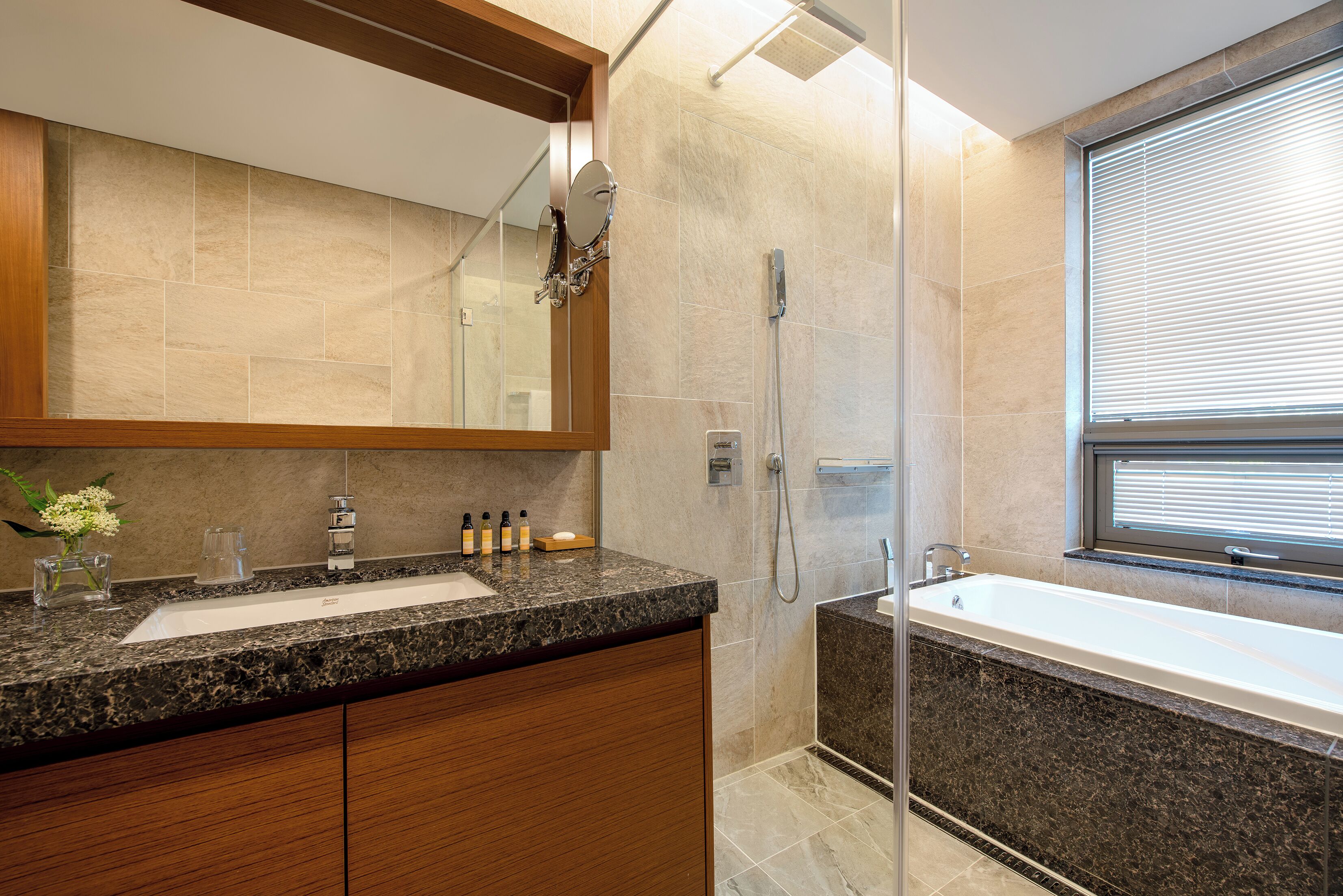 A modern bathroom with a granite countertop, a large mirror, a shower, and a window.