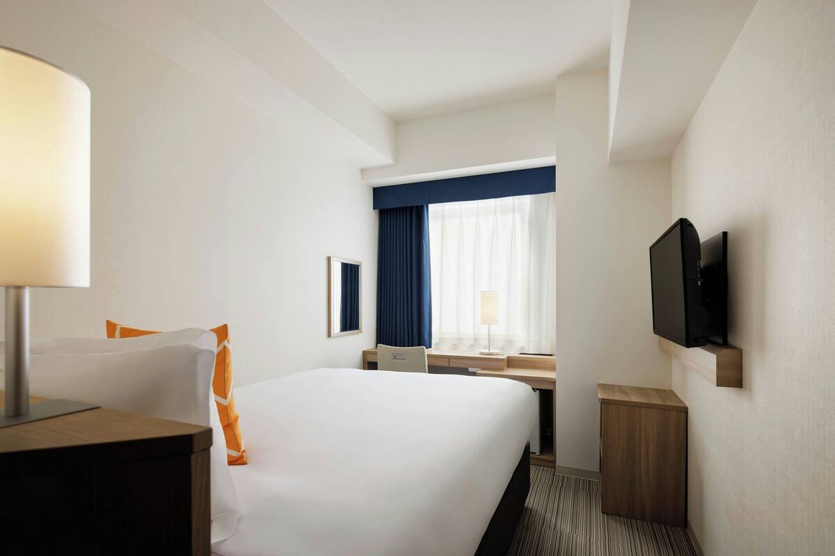 A hotel room with a large bed, a desk, a chair, a TV, and a window with curtains.