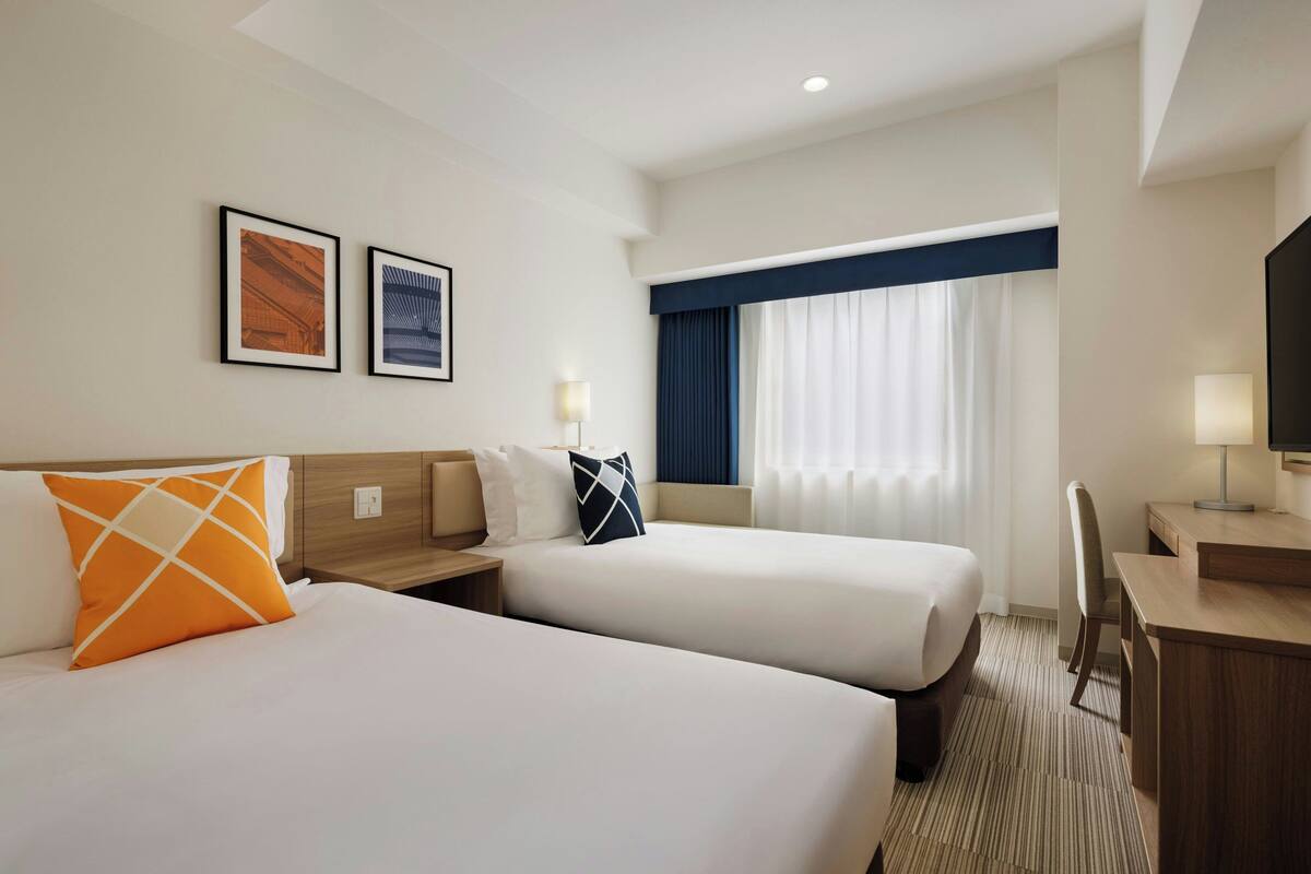 A hotel room with two beds, a desk, a chair, and a window with curtains.