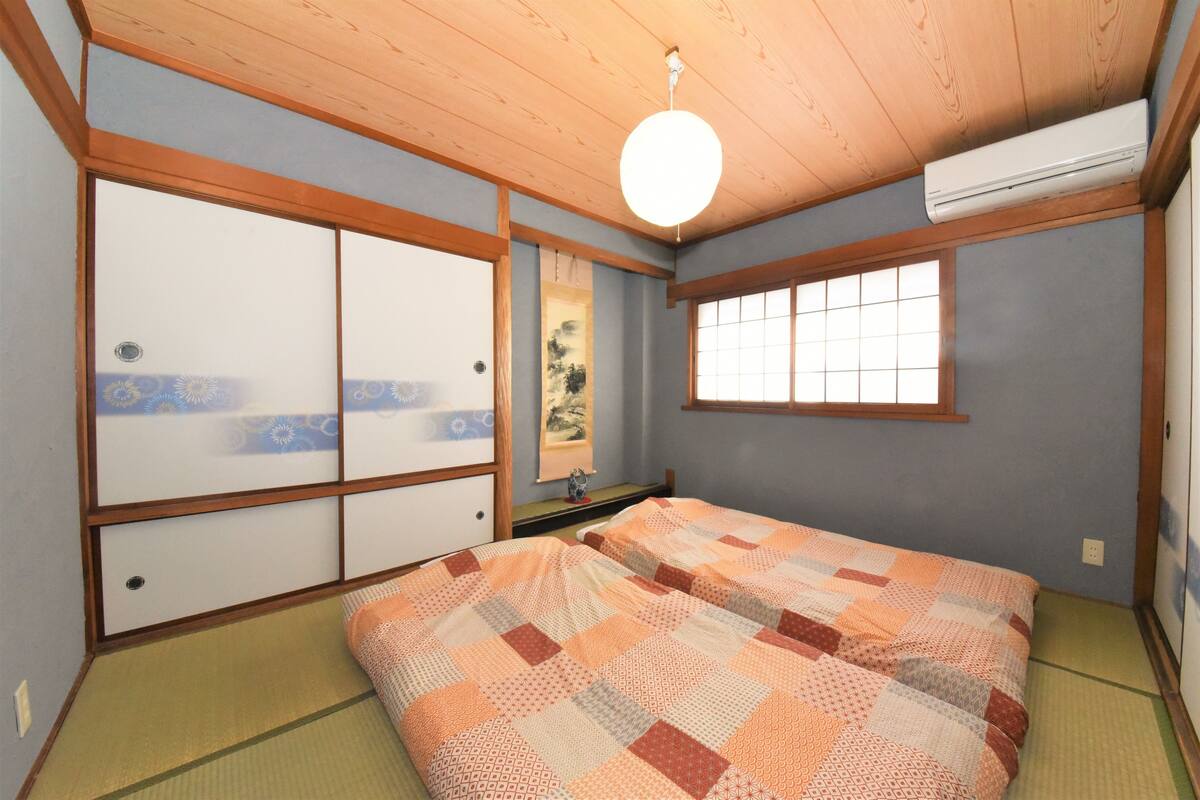 A traditional Japanese room with tatami flooring, a low bed with a patterned blanket, sliding doors, and a ceiling fan.