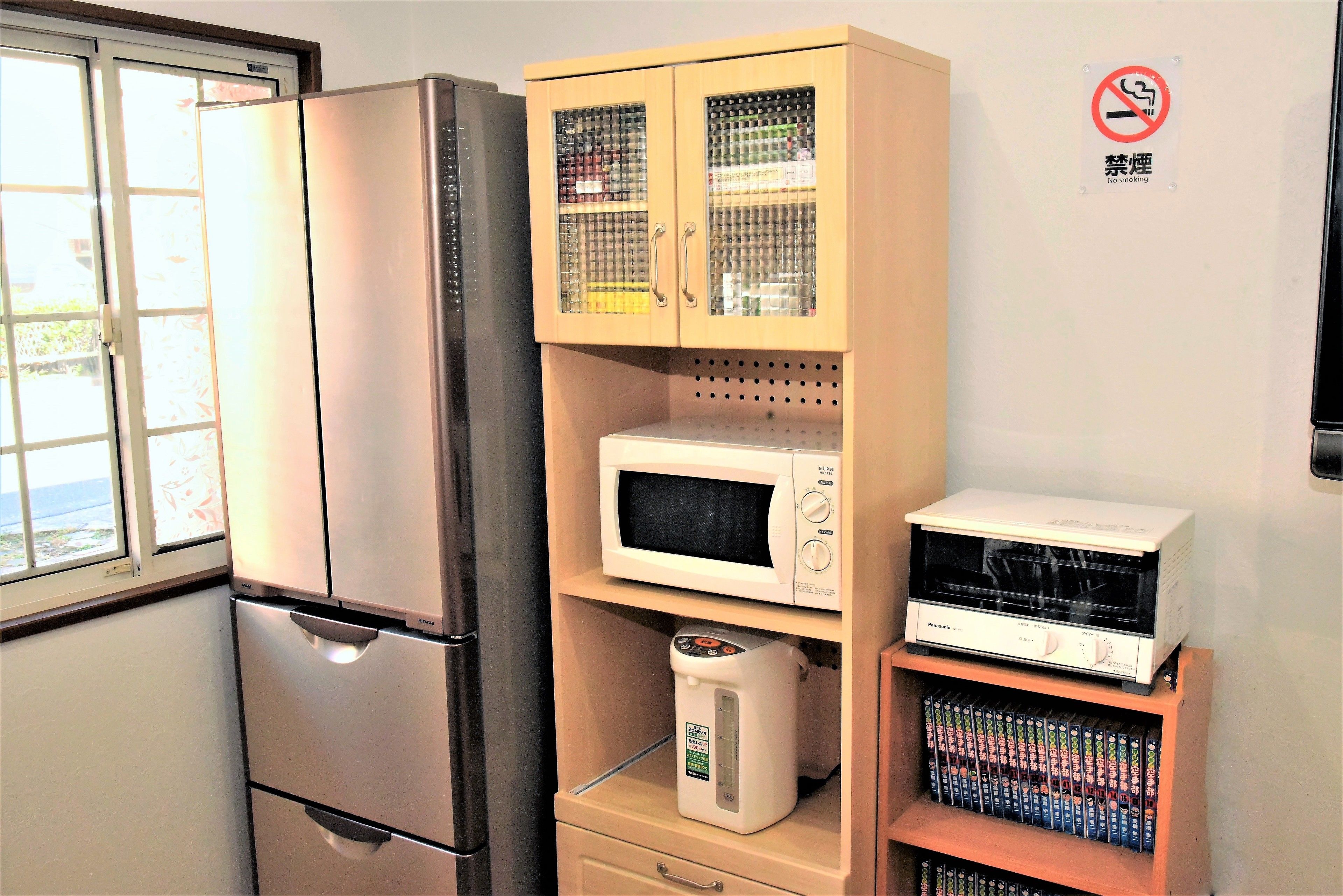 A kitchen with a stainless steel refrigerator, a microwave, a coffee maker, and a shelf with books.