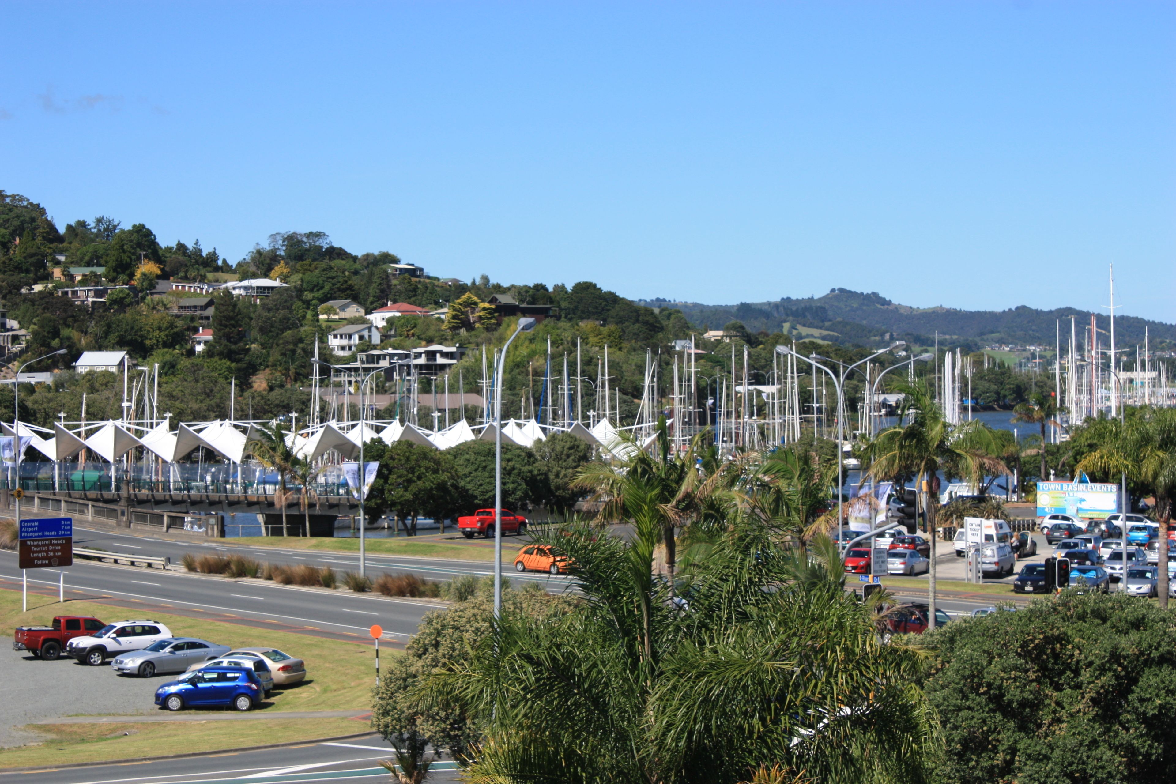 Une route côtière bordée de voitures garées, un port de plaisance avec des voiliers et des maisons situées sur une colline.