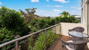 A balcony with a glass table and wicker chairs, surrounded by lush greenery and a clear blue sky.