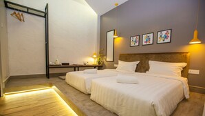 Habitación de hotel con dos camas, piso de madera, mesita de noche, estante y una pared decorada con cuadros.