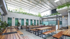 Un café al aire libre con mesas y bancos de madera, un recinto de vidrio con plantas y vista al cielo.