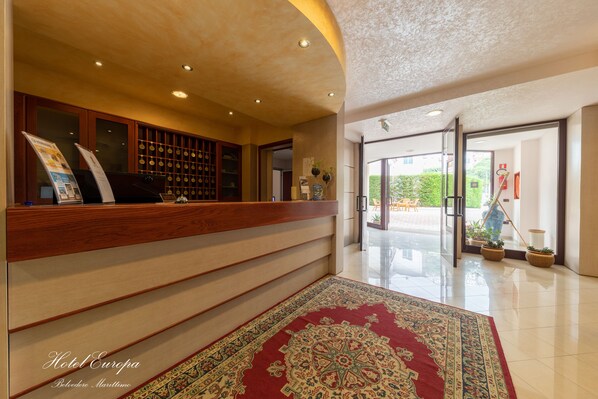 Un hall d’accueil d’hôtel avec un comptoir de réception en bois, un tapis à motifs et une vue sur l’espace extérieur à travers des portes vitrées.