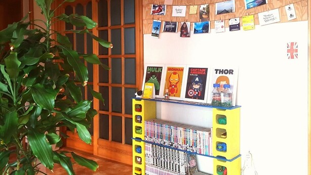 Una habitación con puerta de madera, una estantería con libros y una planta pequeña, y una pared decorada con fotografías y carteles.