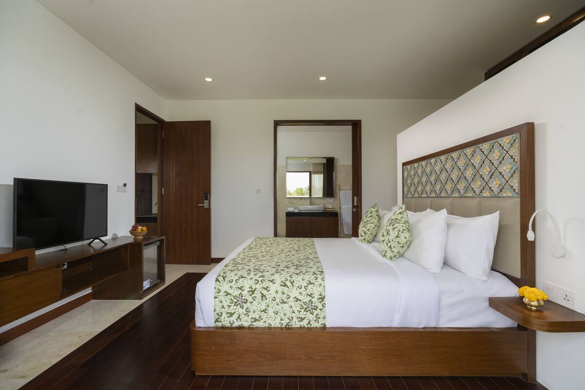 A modern hotel room with a large bed, a TV, a wooden desk, and a bathroom visible through an open door.