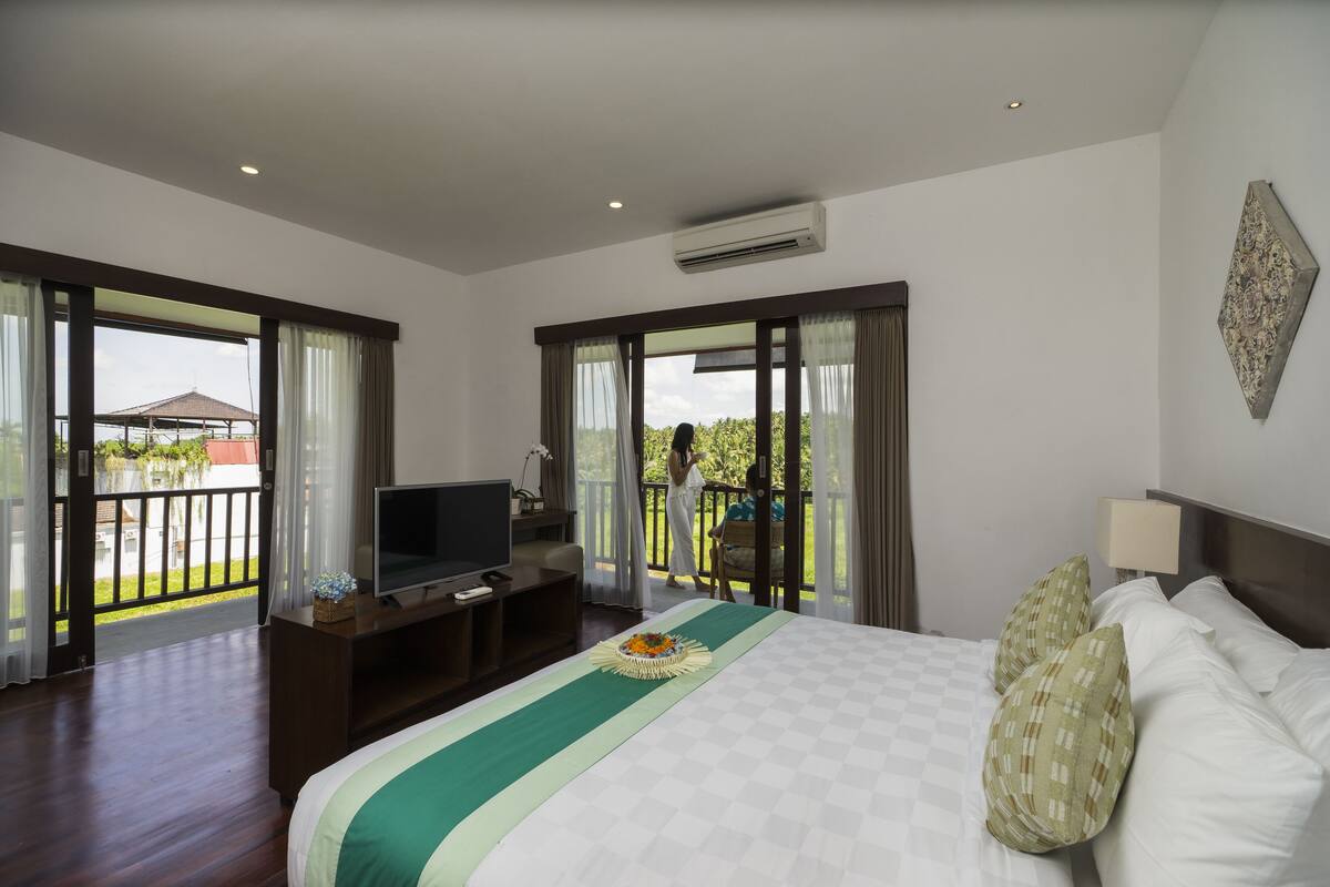 A hotel room with a large bed, a TV, a desk, and a balcony with a view of greenery.