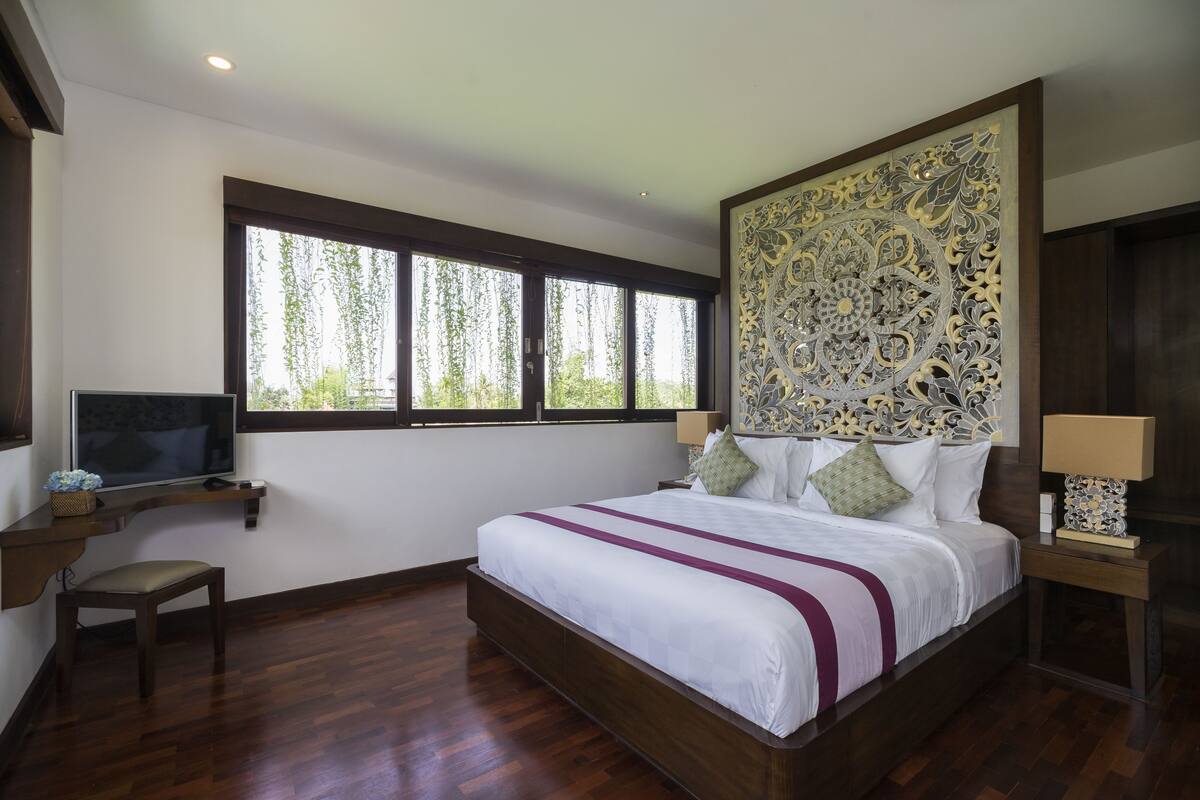A bedroom with a large bed, a TV on a shelf, a window with a view of trees, and a decorative headboard.
