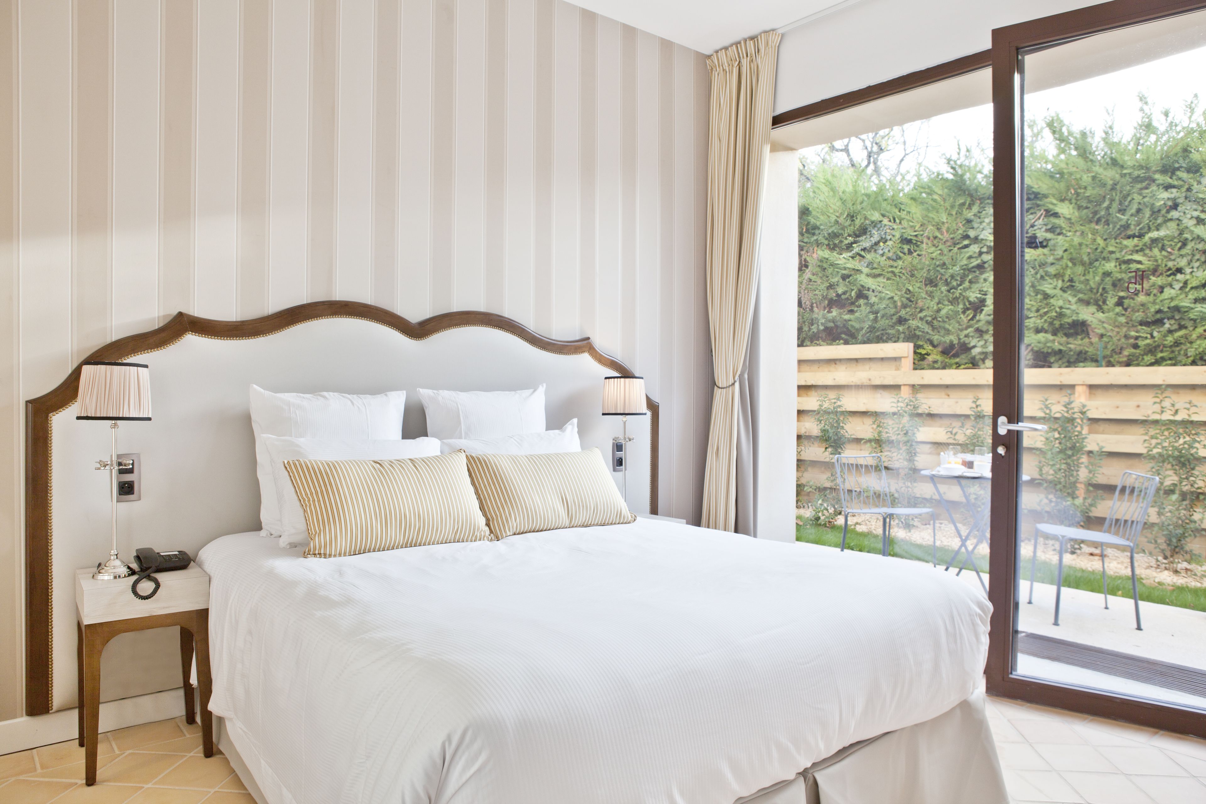 Une chambre d’hôtel avec un grand lit, une table de chevet avec une lampe et une vue sur une terrasse agrémentée de chaises et de plantes.