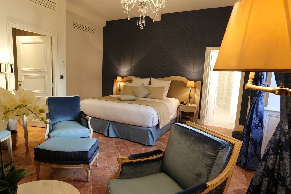 Une chambre d’hôtel avec un grand lit, deux fauteuils, un lustre et une vue sur l’extérieur.
