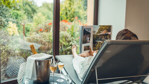 Iemand ontspant in een loungestoel bij een raam met uitzicht op groen, een fles champagne en een glas wijn staan op een dienblad.