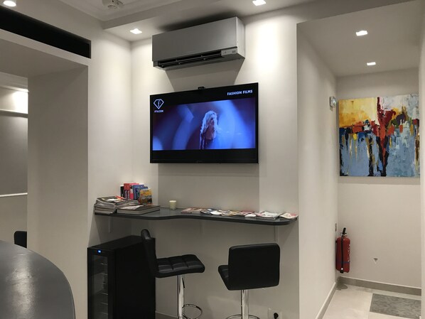 A modern room with a flat-screen TV mounted on the wall displaying 'FASHION FILMS'. There are two black bar stools, a small table with magazines, and a colorful abstract painting on the wall. - Boutique Hotel Neptune Nice (Nice)