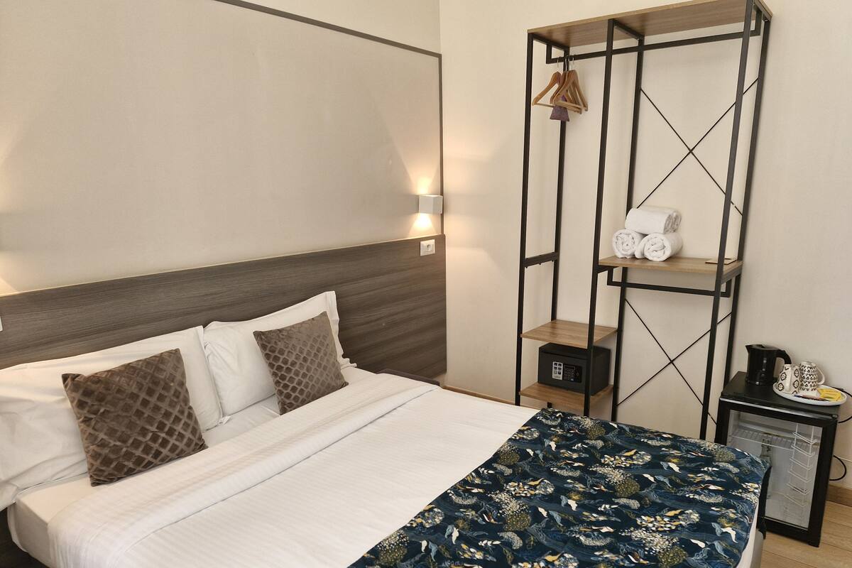 A neatly made bed with a patterned bedspread, a wooden headboard, and a wall-mounted shelf with towels and a kettle.