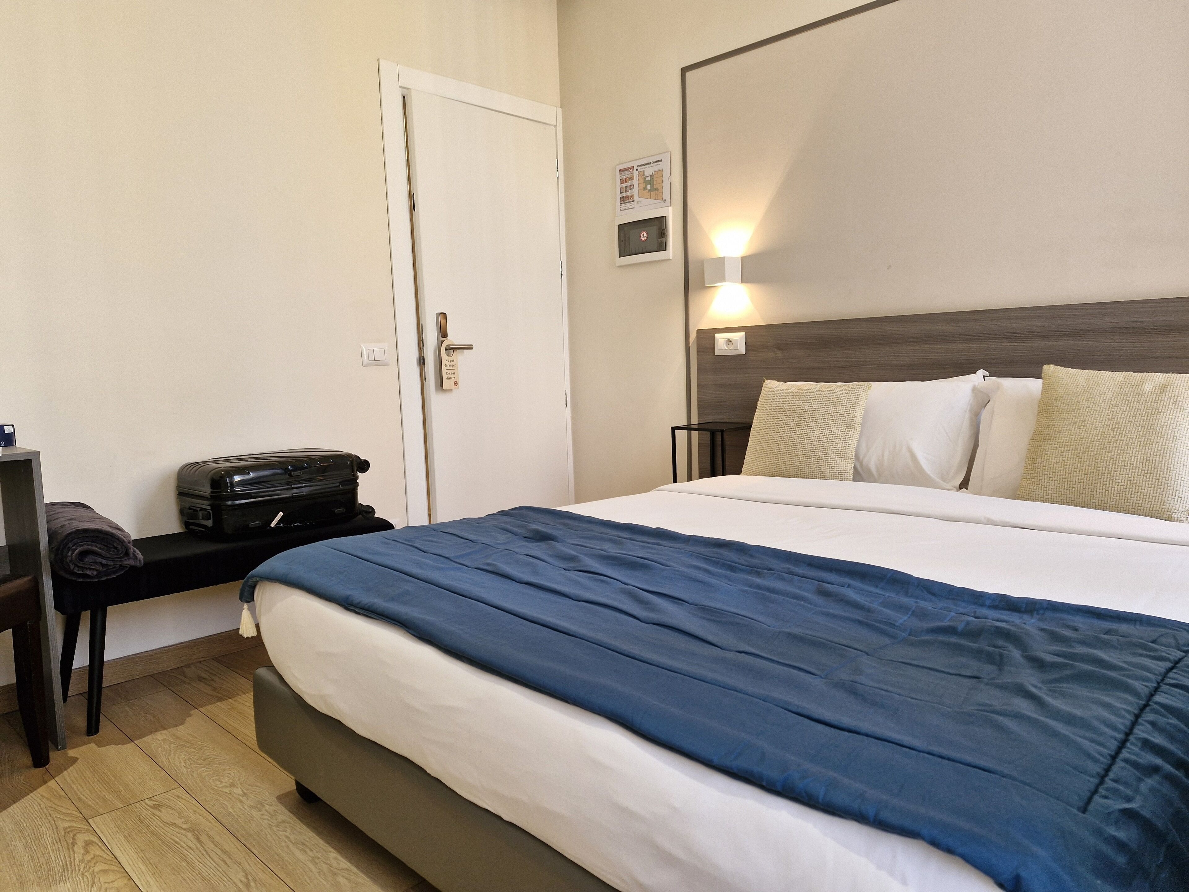 A hotel room with a bed, a desk with a suitcase, a door, and a wall-mounted light.