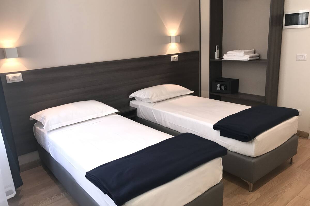 A hotel room with two single beds, a dark headboard, a bedside table with a lamp, and a mirror on the wall.