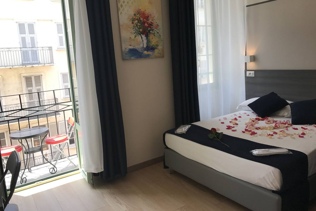 A hotel room with a bed, a painting, a balcony with a table and chairs, and a view of buildings.