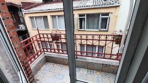 A view from a window overlooking a building with a red metal balcony railing, patterned floor tiles, and a tiled roof.