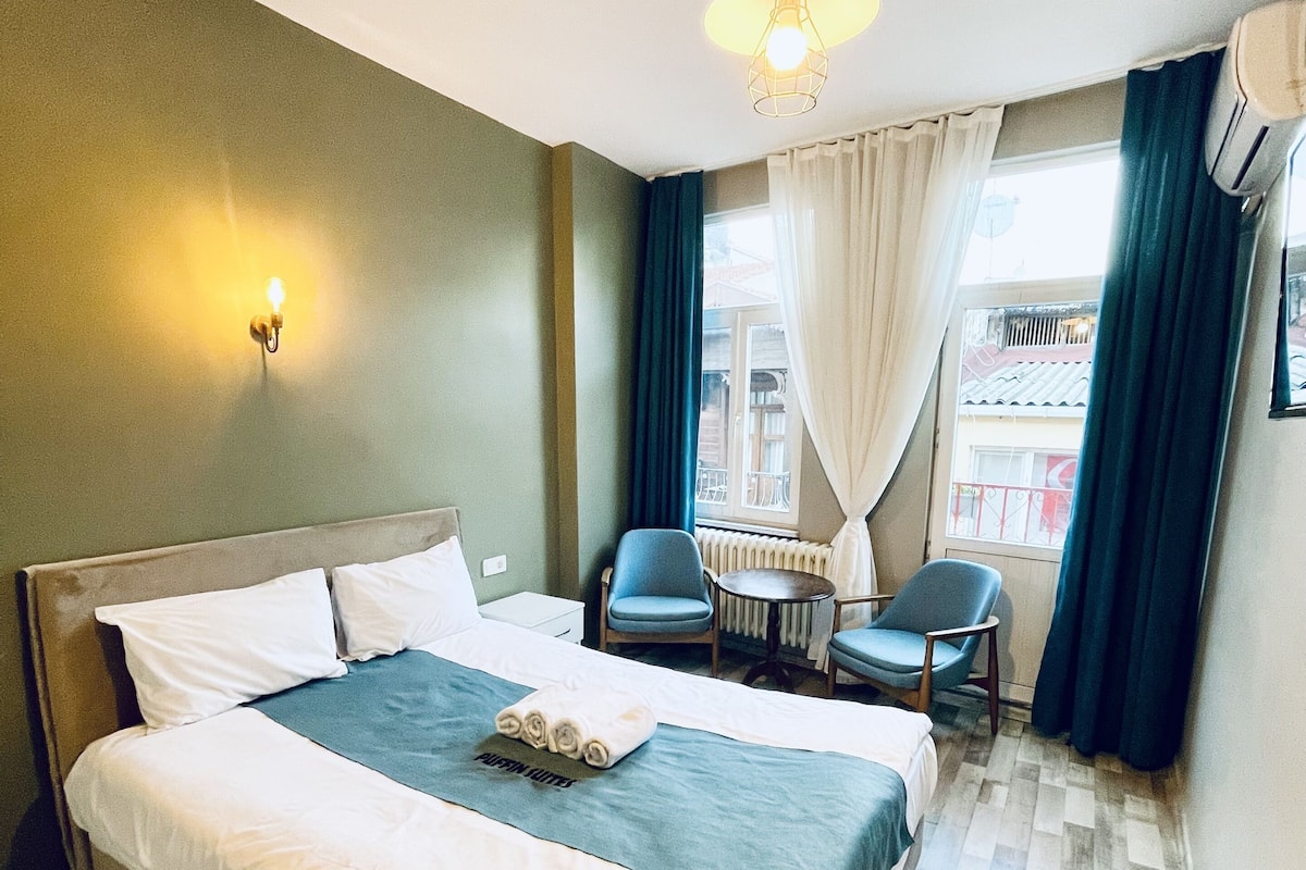 A hotel room with a large bed, a desk, two chairs, a window with curtains, and a hanging light fixture.