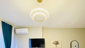 A room with a black chair, a small table with a chair, a mounted TV, a wall-mounted air conditioner, and a ceiling light fixture.