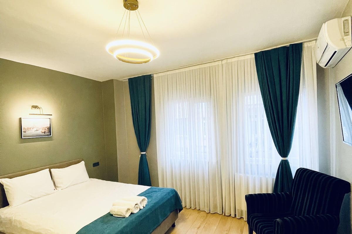 A hotel room with a bed, a chair, a ceiling light, a wall-mounted air conditioner, and a window with curtains.